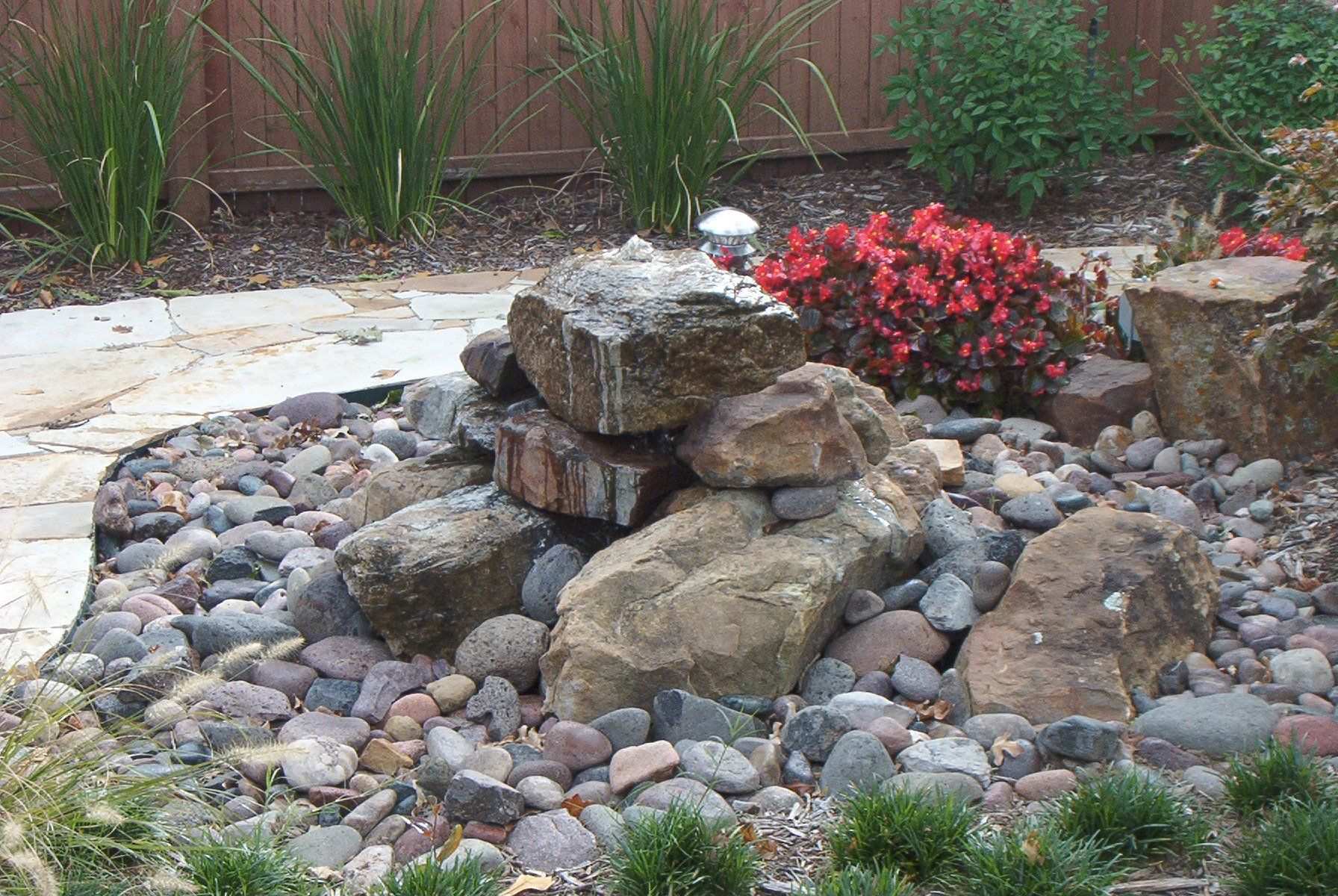 Boulder water feature set in river rock with red flowering shrubs behind
